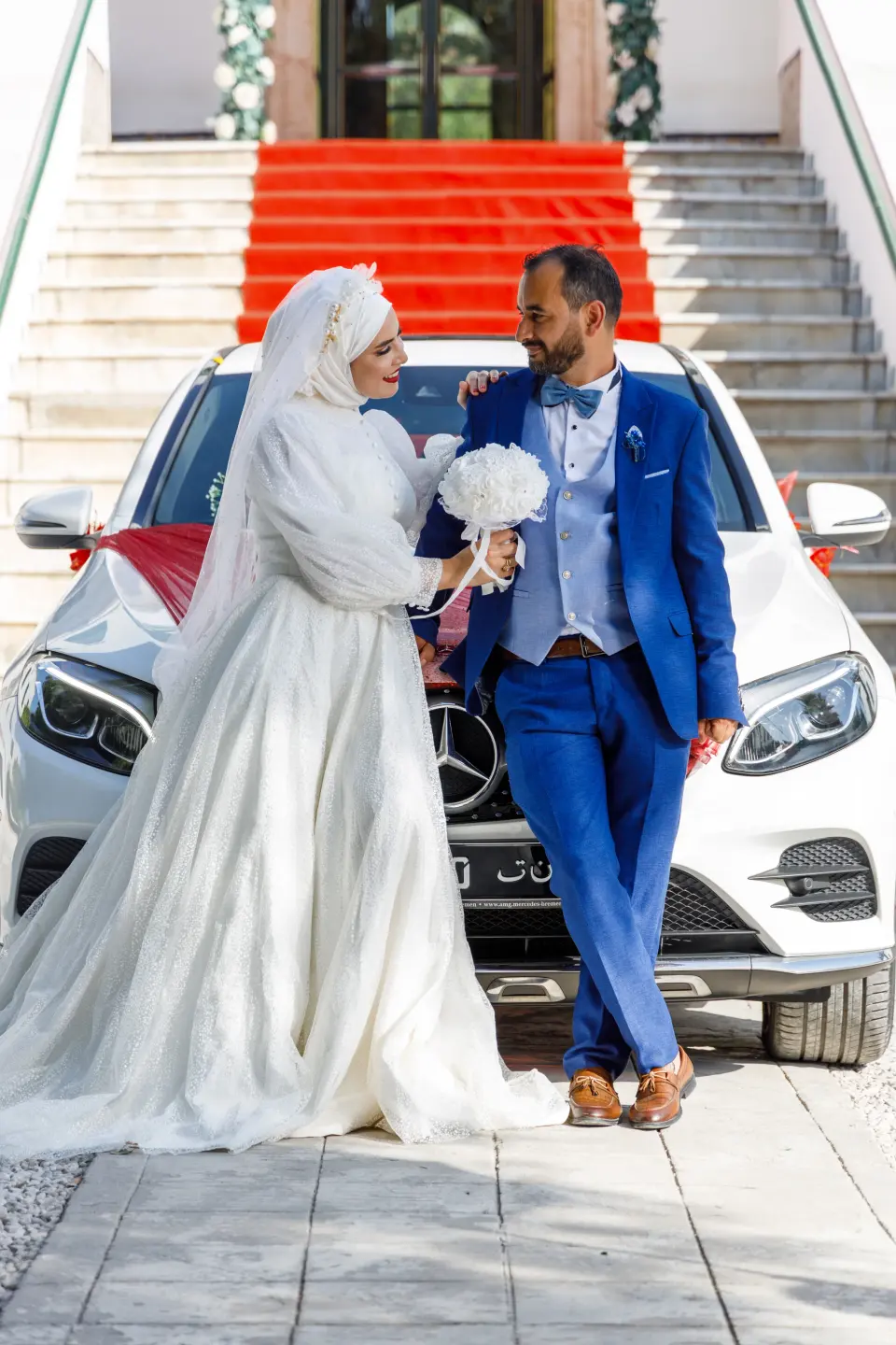 Mariés devant une voiture blanche au pied d’un escalier au tapis rouge